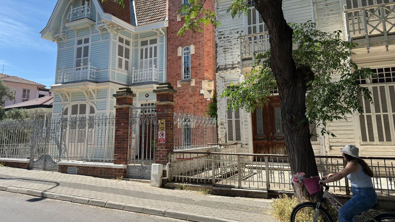 A light-blue wooden Büyükada mansion with a brick tower behind an iron fence, as a woman cycles past on the car-free street.